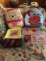 Overview of Pooh and Snoopy backpacks, framed cross-stitch, pouch, mouse pad, and book arranged on floral couch.