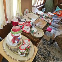 Overall view of Christmas themed dishware and serving pieces arranged on a wooden cabinet, including plates, mugs, trays, and large snowman cookie jar.