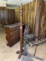 Full view of tall wooden coat rack with four-legged base and multiple wooden pegs, standing on concrete floor in basement setting.