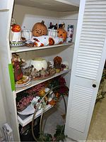 Three-shelf cupboard with assorted fall, Halloween, and Thanksgiving decorations showing ceramic pumpkins, floral arrangements, and plates.
