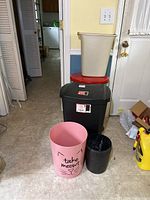 Five garbage cans arranged indoors on tile floor near a door. Includes pink cat-themed can, small black textured can, beige step-on can, red step-on can, and large black rectangular step-on can.
