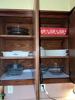 Wooden cabinet shelves containing white Corning Ware casserole with blue floral pattern, various clear glass pie plates and bowls, red Pioneer Woman casserole dish with colorful floral and bird design.