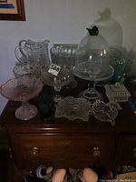 Photo showing multiple clear and colored glass serving ware pieces arranged on a wooden surface, including pitchers, compotes, candy dishes, decorative vase, and covered dome with bird finial.