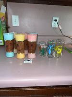 Full lot of eight glasses on kitchen counter, showing Siesta Ware tiki glasses and Flintstones-themed glasses.