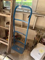 Full view of blue metal hand truck standing upright in a garage near a ladder and various paint cans.
