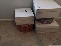 Four hat boxes stacked on floor: two plain white boxes, one floral patterned box, and one smaller white box with label.