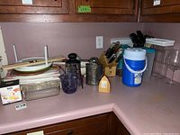 Photo showing multiple kitchen items including cutting boards, various kitchen utensils including knives in block, plastic containers, vintage sifter and egg beater, and ice bucket cooler.