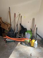 Wide view showing assortment of lawn care tools and supplies including rakes, shovels, bags, and containers against garage wall.