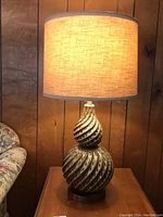 Photo of the lit Mid Century Modern table lamp showing the woven look of the shade and warm light output, lamp placed on wood table with wood paneled background.