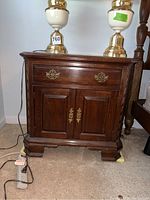 Front view of one wooden nightstand showing dark wood finish, brass hardware, single drawer above two-door cabinet, and bracket feet.