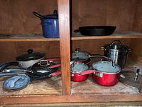 Large black cast iron skillet, blue enamelware pot, multiple red and black pots and pans with glass lids, stainless steel double boiler in wooden shelving
