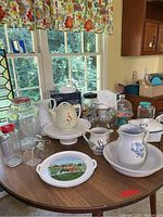 Photo showing multiple beverage containers, pitchers, and serving items arranged on a round wooden table near a window with floral curtain.