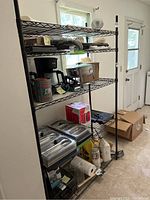 Front view of the black metal utility shelf showing multiple kitchen items including a coffee maker, metal pans, containers, paper towels, and other miscellaneous items
