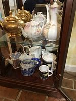 Shelf inside wooden cabinet holding multiple tea pots and pitchers including gold, porcelain with ruffled edges and floral designs, ceramic pitcher, and mugs.