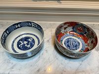 Pair of Asian porcelain bowls on white marble surface showing interior and exterior patterns, one blue and white, one red floral with gold edge.