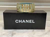 Front view of turquoise resin cuff bracelet with gold leaf stripes and pearl CC logo displayed atop black Chanel box