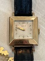 Close-up front view of square watch face showing silver-tone dial, Marvin logo, gold-tone hands and hour markers.