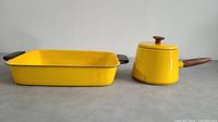Yellow rectangular cast iron serving tray with black handles and yellow Michael Lax designed cooking pot with lid and wooden handle side by side