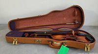 Image of violin inside open brown hard case with purple interior lining and attached bow.