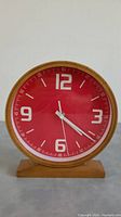 Front view of vintage battery operated clock with red face, white numerals, and wood frame and base.