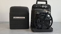 Front view of Bell & Howell Model 256 projector with attached film reel and carrying case placed next to it.