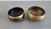 Two Tibetan brass bowls showing exterior and interior designs side by side on a flat surface.