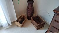 Photo showing the tall woven vase flanked by two woven baskets of different sizes on carpeted floor beside wooden furniture.