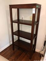 Front view of a wooden three-shelf bookcase with a dark brown finish standing on a hardwood floor against a white wall.
