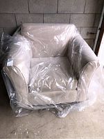 Beige fabric armchair covered in clear plastic, showing overall size and condition.
