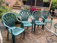 Four green plastic patio chairs arranged outdoors on a brick patio near glass outdoor table.