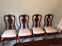 Photo showing four wooden dining chairs in a row with white fabric seats and carved backs.