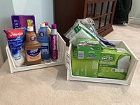Two white plastic bins on carpet containing cleaning supplies. Various bottles and refill packages visible including Swiffer and Vileda brands.