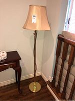 Full view of the floor lamp showing the brass-colored base, curved slender stand, beige fabric lampshade, and power cord on the floor.