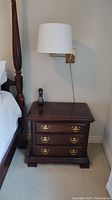 Full frontal view of the Kincaid wooden night stand placed next to a bed with a wall lamp and phone on top showing condition and style.