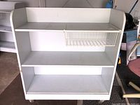 Front view of white painted wood rolling cart with three open shelves and wire basket under middle shelf. Shows wear on surfaces and rolling casters.