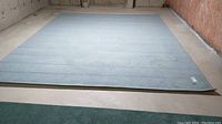 Full view of the large light blue/gray machine made rug on a basement floor, showing the full area and pattern.