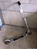Full view of silver kick scooter leaning against a concrete wall, showing handlebars, deck, and wheels.