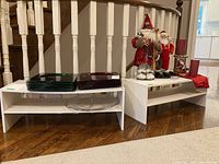 Pair of white melamine shoe stands placed on hardwood floor in front of a staircase railing, one holding green and red plates, both showing scratches.