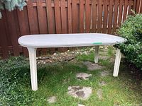 Wide angle photograph of the large beige plastic patio table positioned outdoors on grass near wooden fence and bushes.