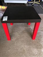 Square table with black top and red legs photographed from an angled front view showing wear on the finish and solid construction.
