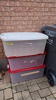 Three plastic storage totes stacked outside against a brick wall next to a black waste bin. The totes have wear, with the top tote having a white lid, the middle a red lid, and bottom a grey lid. Visible scratches and tape residue on the totes.
