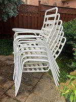 Six white metal patio chairs stacked outdoors on a stone patio, showing design and condition of chair frames