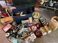 Overview of various Christmas decorations and items on floor and in plastic bin, includes tins, napkins, ornaments, boxed angel figurines, and heart-shaped dishes.