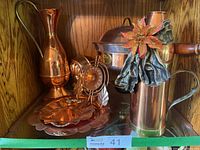 Shelf display with shiny copper serving and kitchen items including chafing dish, urns, plates, and copper thermometer.