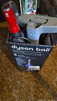 Opened Dyson Ball box showing vacuum parts inside including the red handle and grey plastic parts