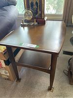 Side view of one wooden mid century modern side table showing top surface, legs with brass feet, and lower shelf