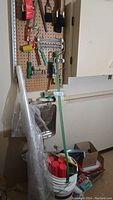 Wide view showing pegboard with hanging paint brushes, roller frames and rollers, roller trays, glue spreading trowels, plastic sheeting roll, bucket with red capped containers and other painting tools.