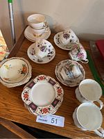 Multiple teacups, saucers, snack plates, and bowls arranged on wooden surface showing variety of floral and scenic patterns.
