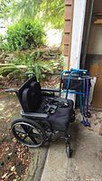 Side view of black Breezy 600 wheelchair with padded armrests and folding walker behind it, set outdoors near a garage