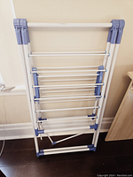 Full view of the foldable drying rack standing upright and folded flat, showing multiple horizontal metal rods and blue plastic hinges